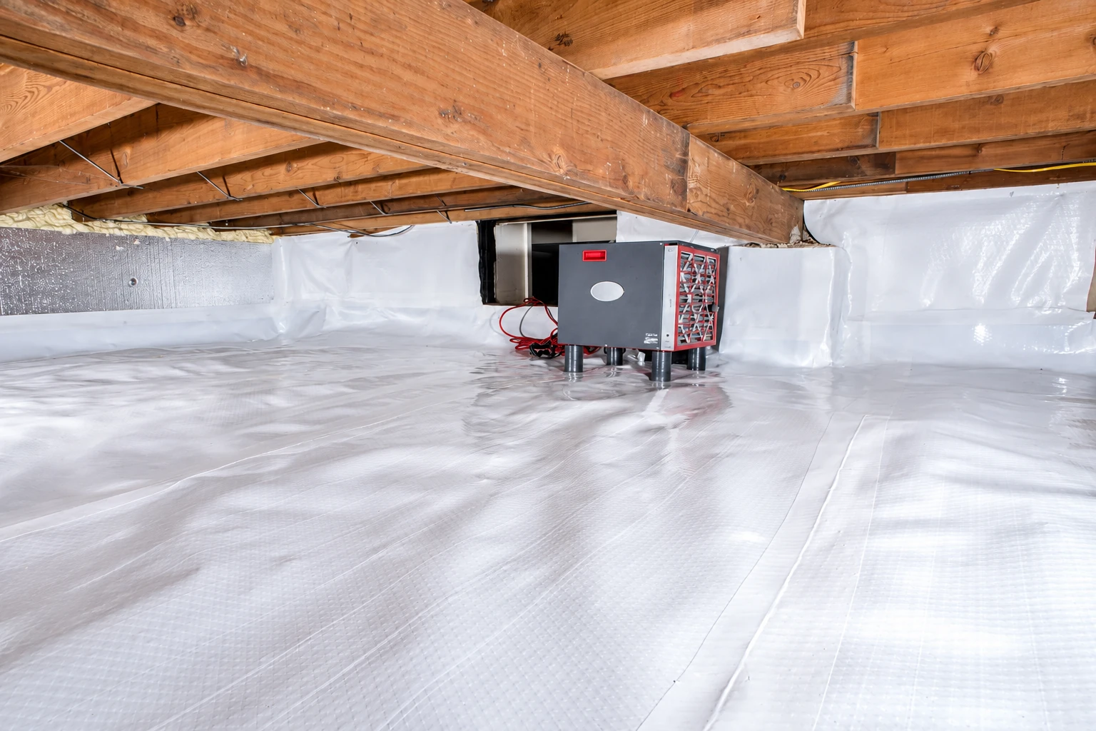 A low crawl space has a white plastic floor and wall liner beneath exposed wooden joists, with a gray equipment unit near the back.