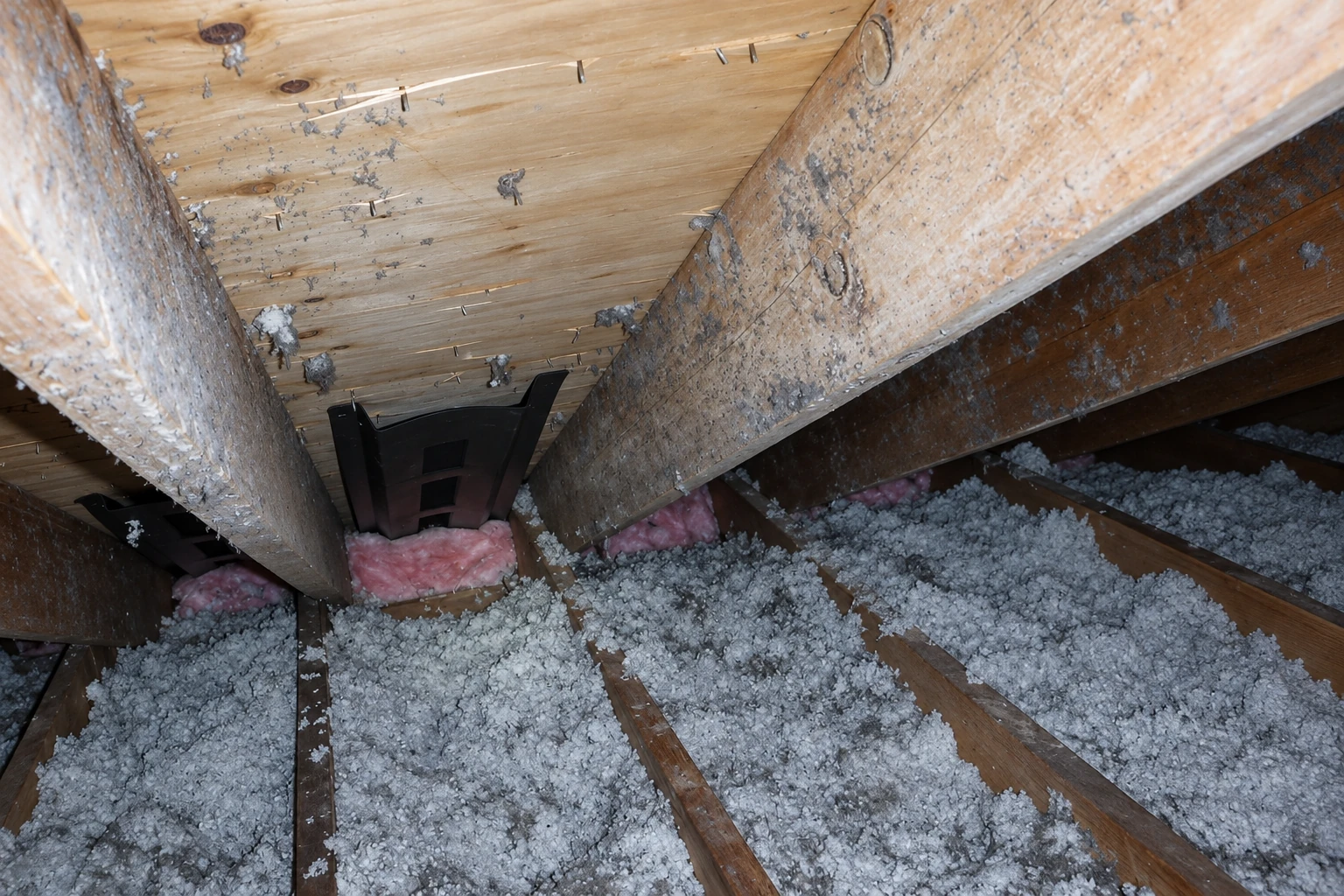 Attic floor with loose gray insulation between exposed wooden joists and pink batts around a roof vent.