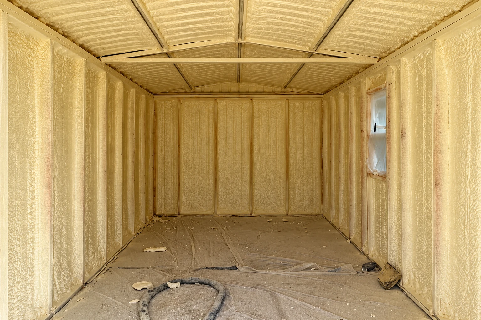 Commercial space with yellow spray foam insulation covering the walls and ceiling; a tarp and hose lie on the floor.