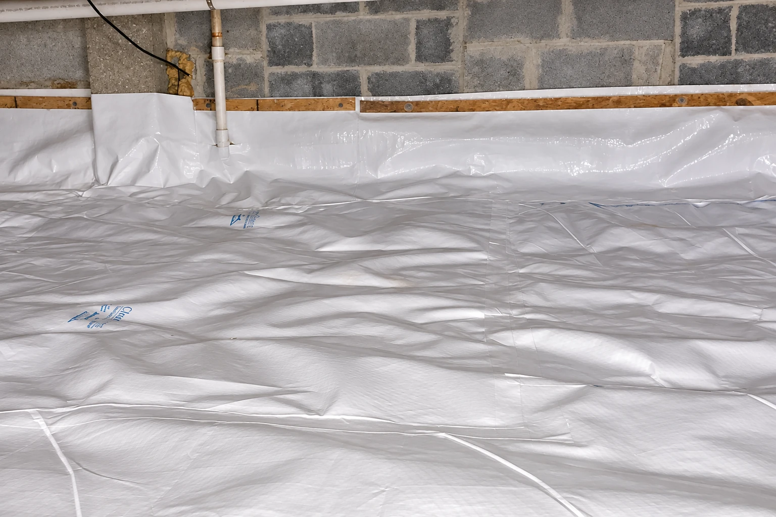 Crawl space floor and lower block walls are covered with a wrinkled white vapor barrier beside exposed pipes and wiring.