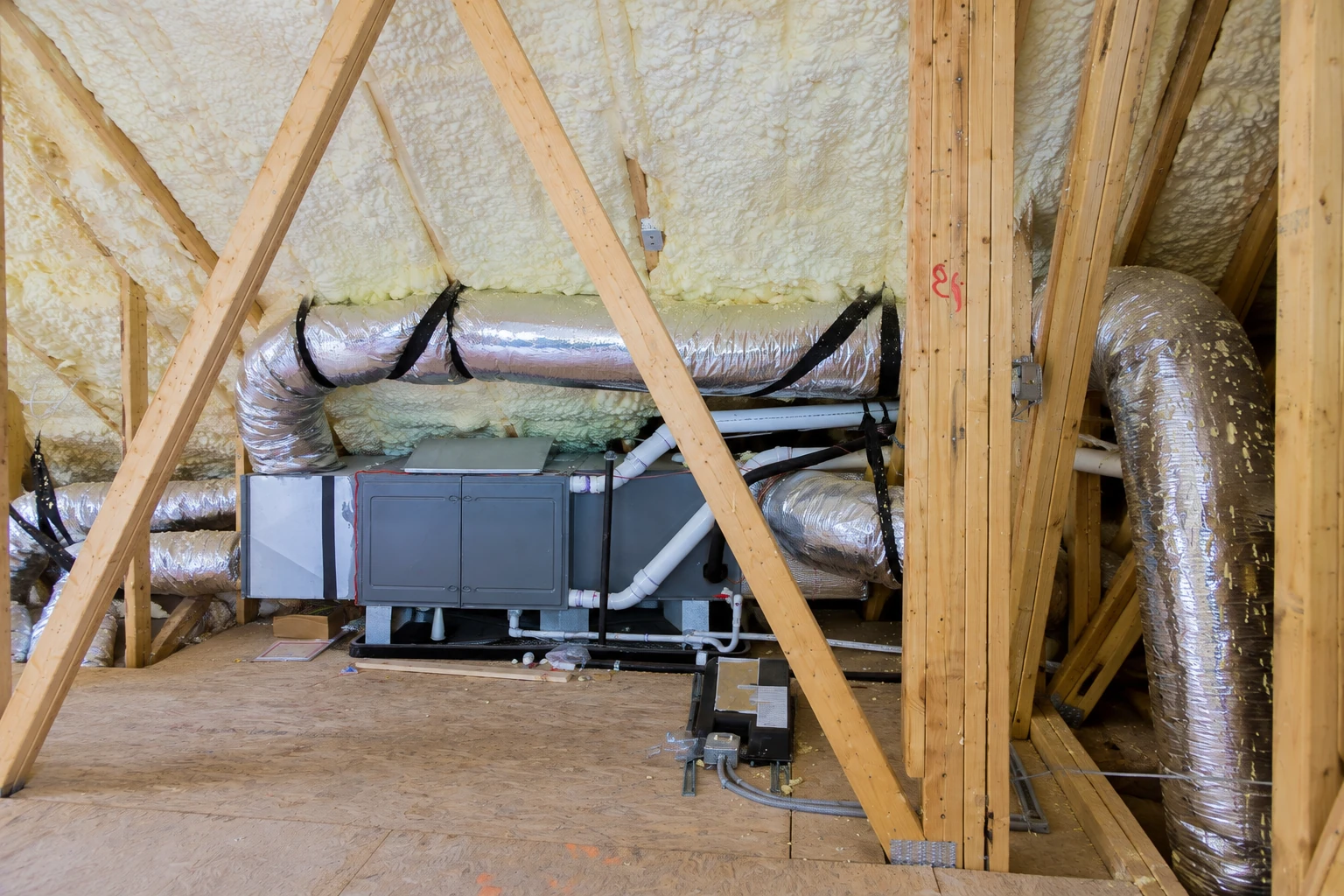 An attic HVAC unit and silver ducts sit beneath roof trusses coated with pale spray foam insulation.