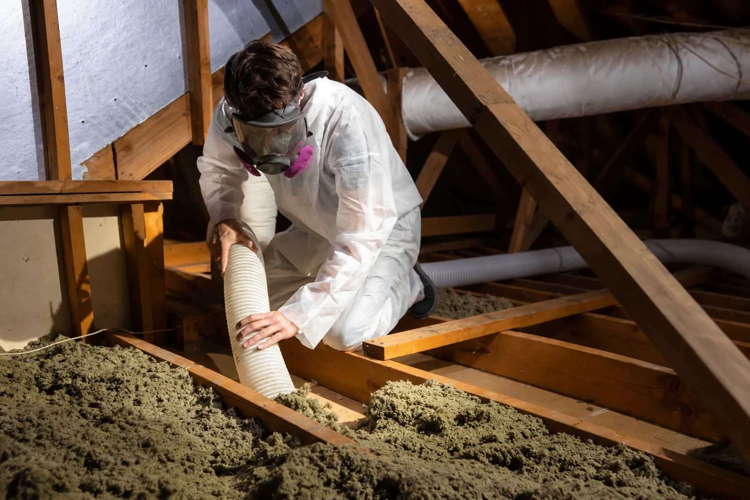 A worker in protective coveralls and a respirator vacuums loose attic insulation through a flexible hose.