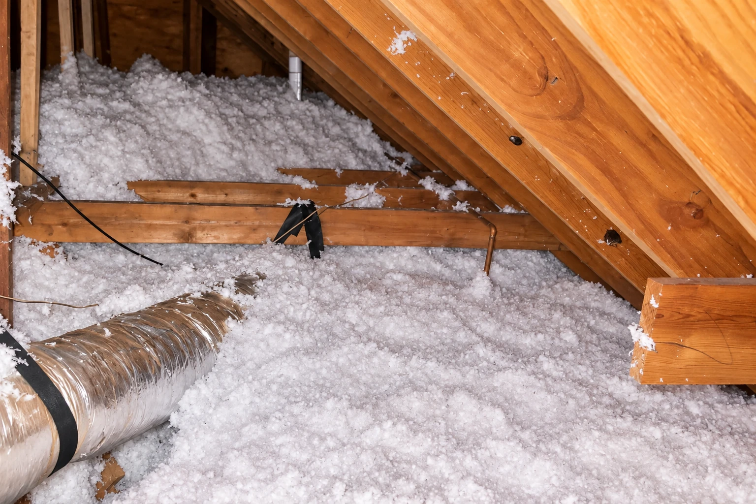 Attic with loose white blown-in insulation spread across the floor between exposed wooden rafters and a duct.