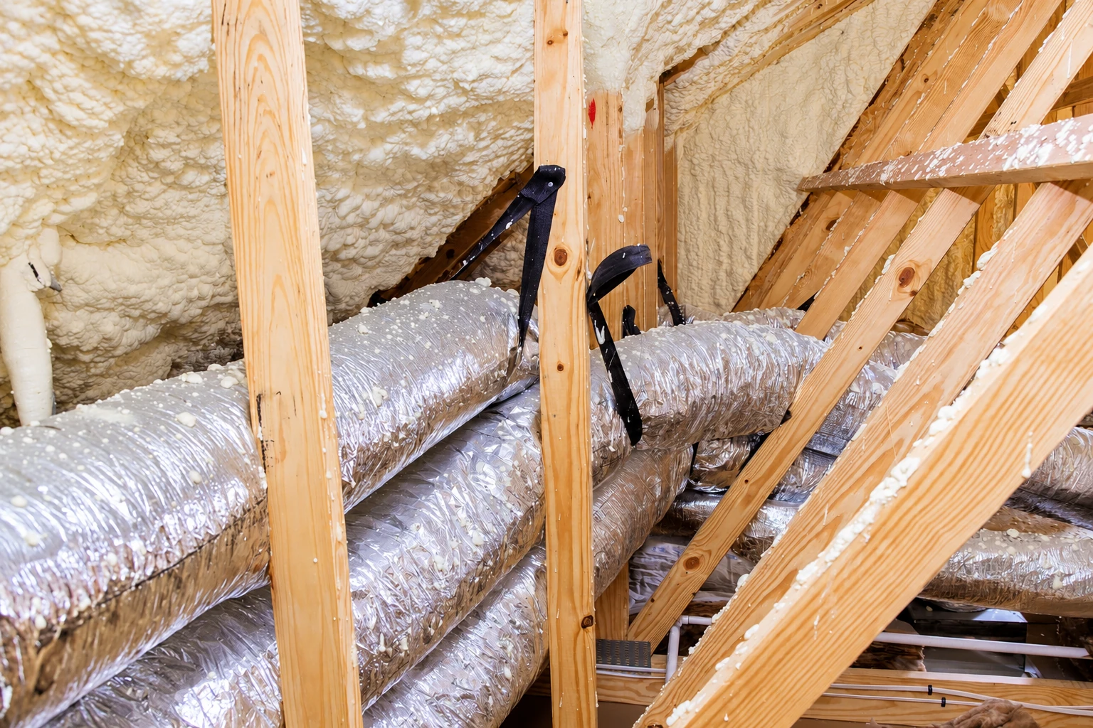 Spray foam insulation coats an attic roof between exposed wooden rafters above silver flexible ducts.