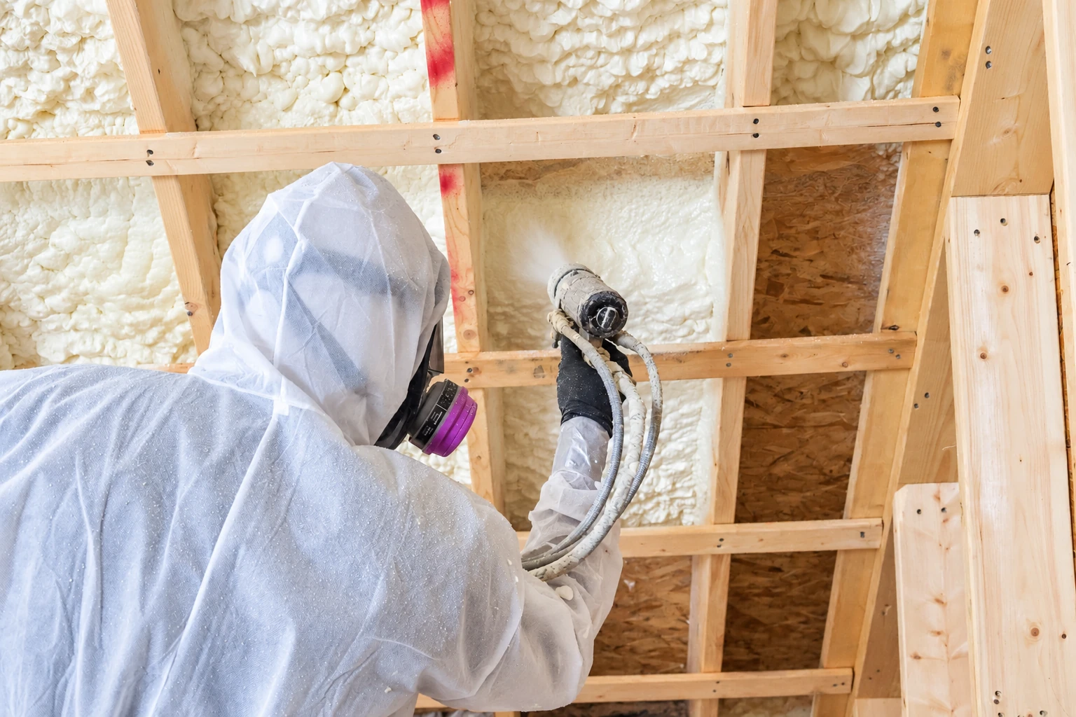 Worker in protective gear sprays foam insulation into open wall framing, with foam already filling adjacent cavities.