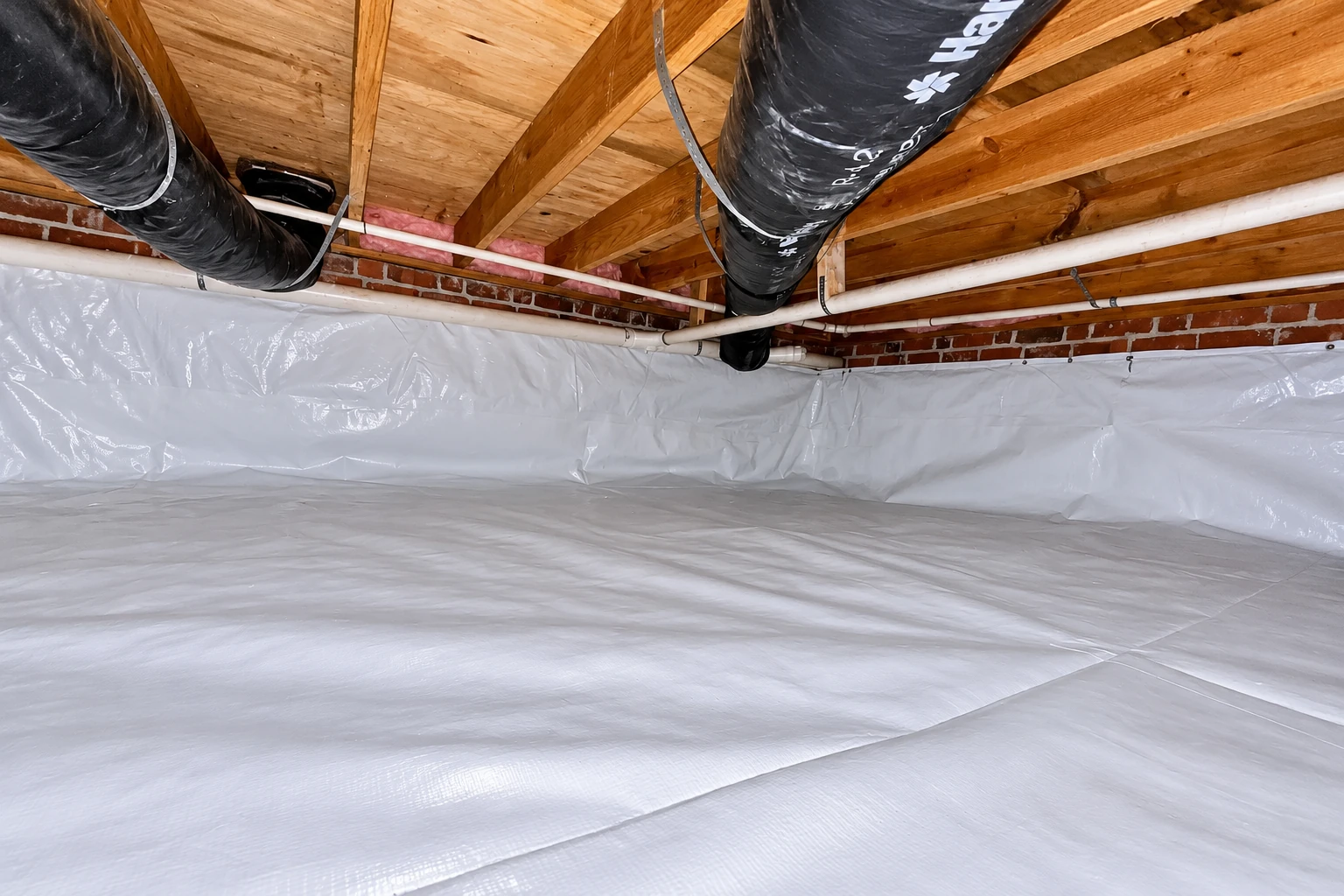 White plastic vapor barrier covers a crawl space floor and lower walls beneath exposed joists, ducts, and pipes.
