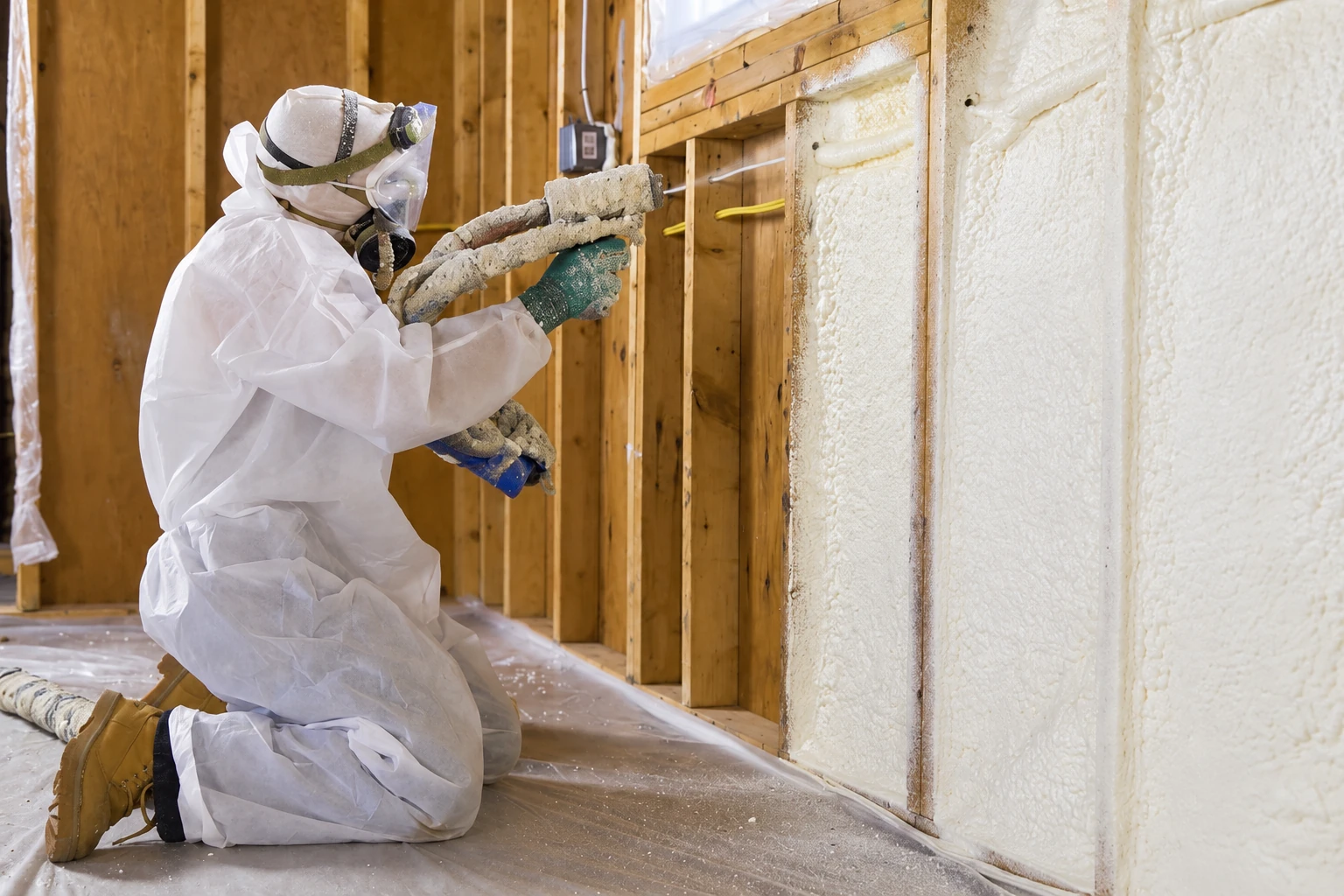 A worker in protective gear sprays foam insulation into an open wall cavity between wooden studs.